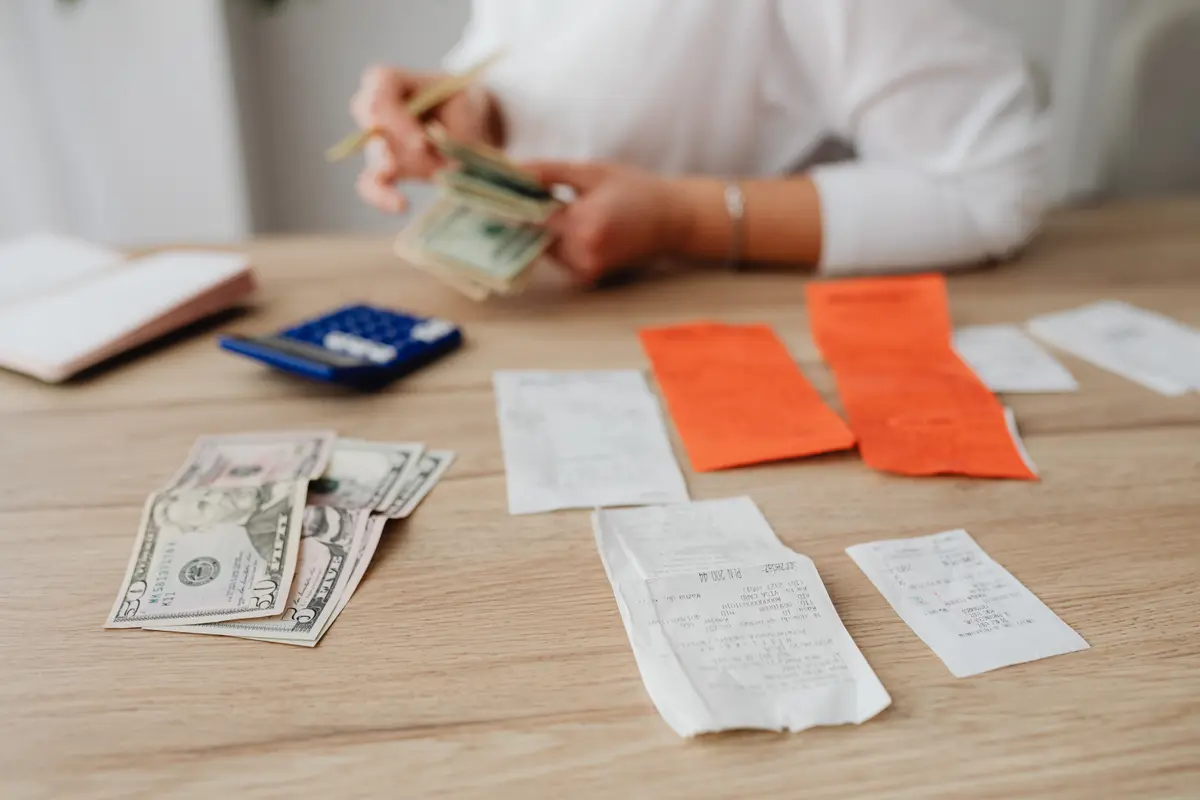 Desk with dollar bills, paper receipts and a calculator, someone counting money in the background