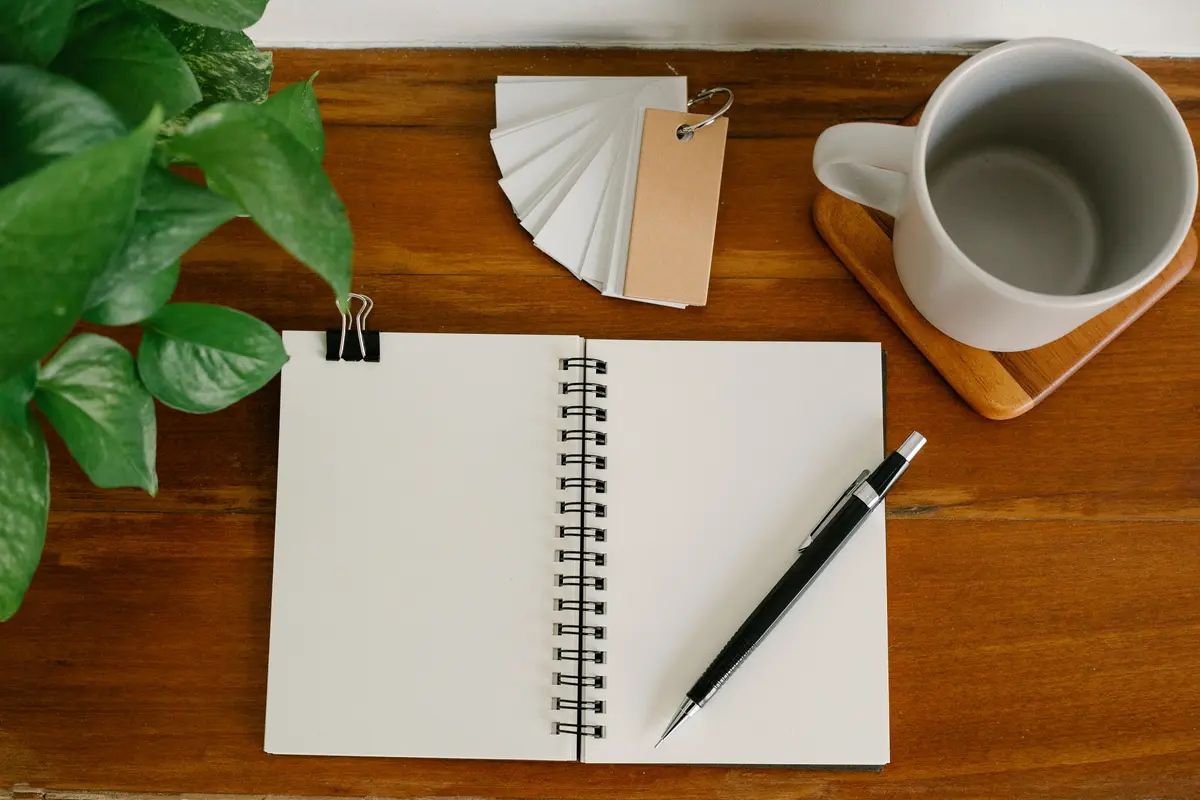Open spiral notebook with a pencil on a wooden desk next to a coffee mug and a plant