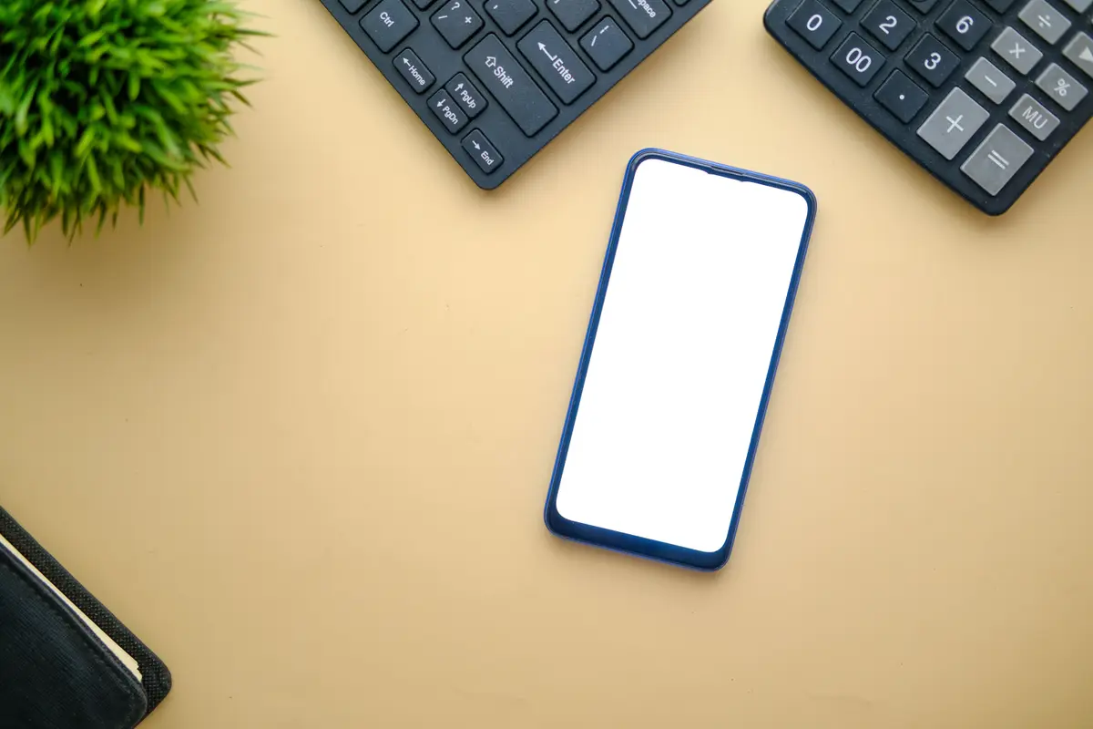 Smartphone with a blank white screen lying on a yellow desk between two keyboards