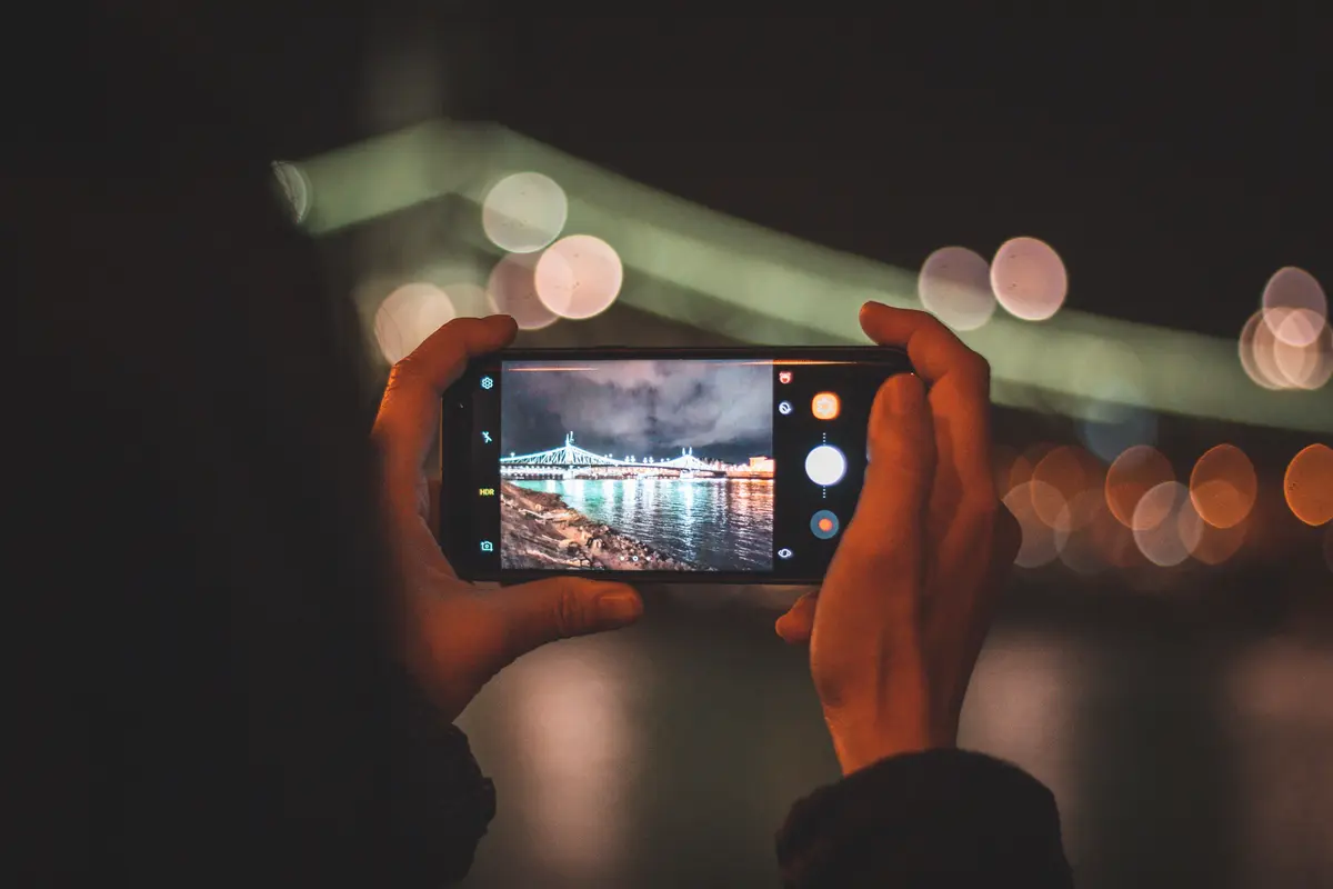 Two hands holding a phone sideways to photograph a city bridge lit up at night