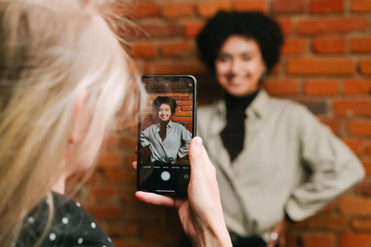 Someone photographing a smiling woman against a brick wall with a phone