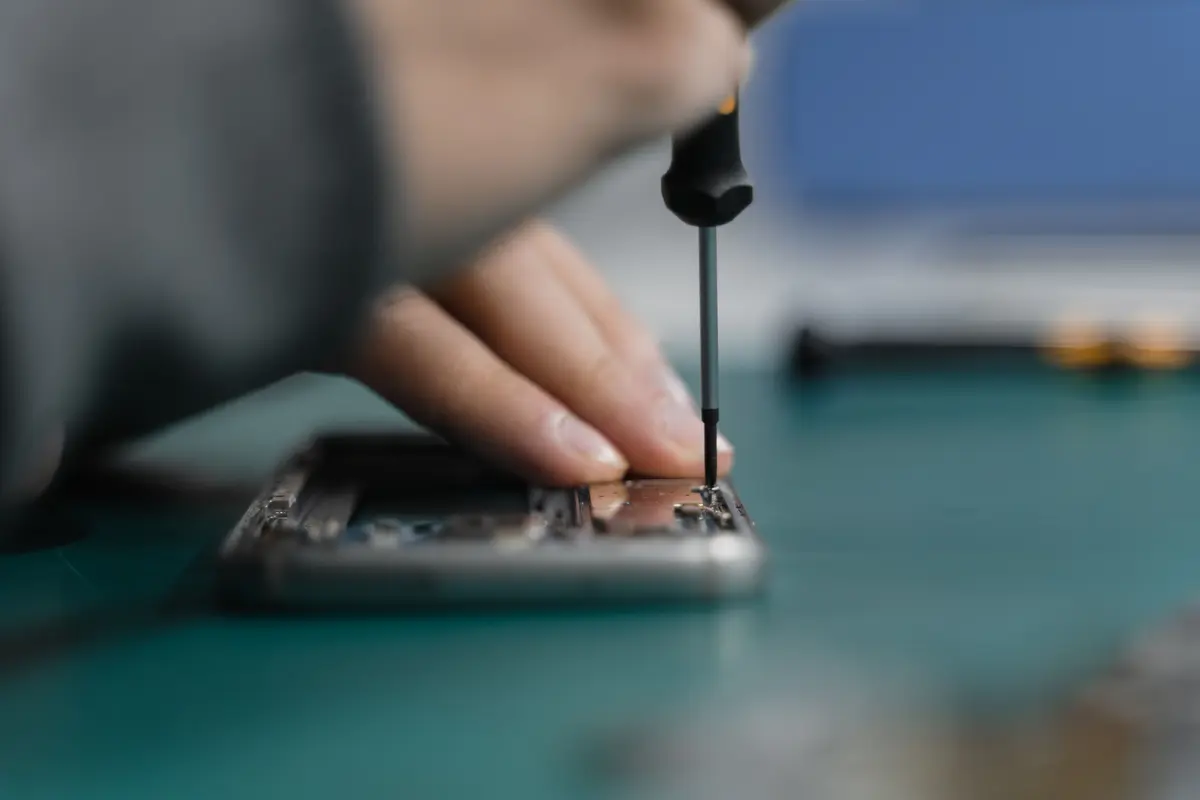 A technician's hand holding a precision screwdriver over an opened phone on a green repair mat